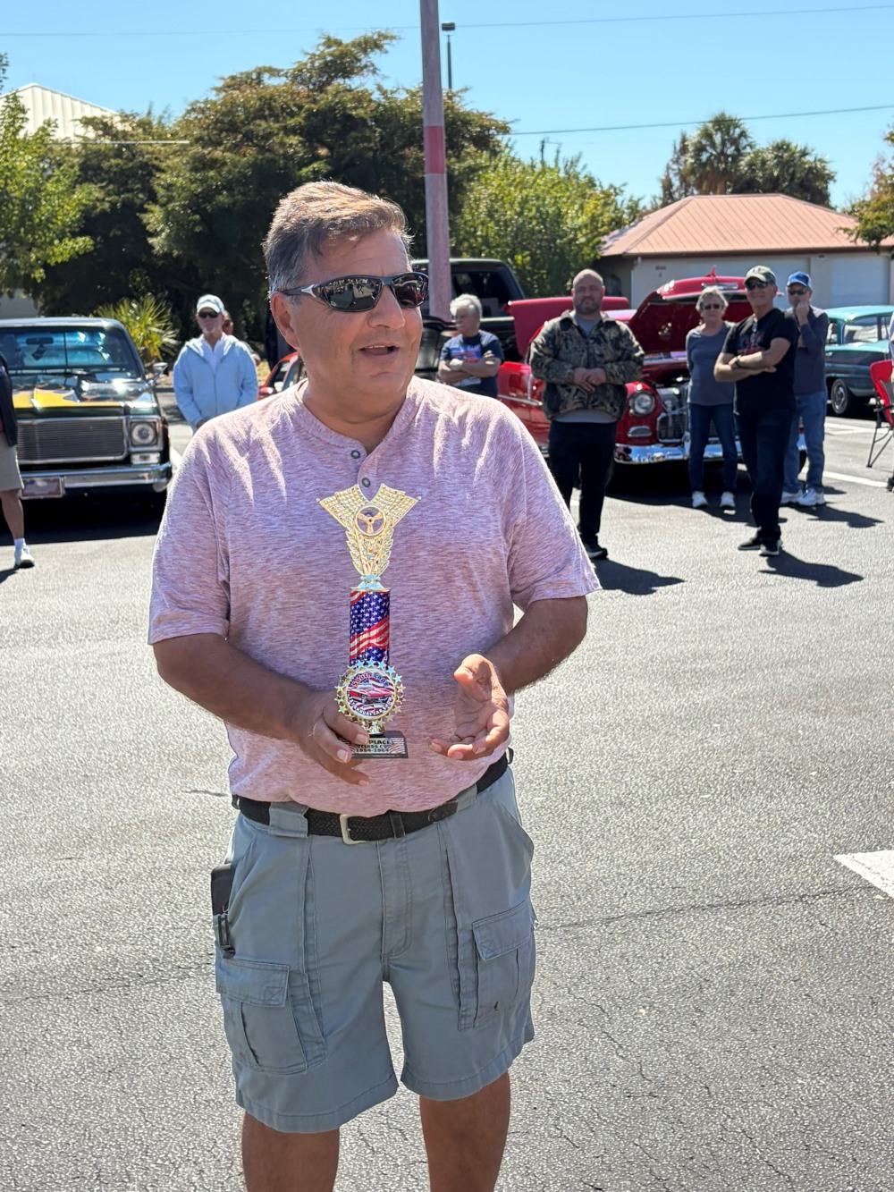 Man in a pink shirt and cargo shorts holding a red, white, and blue car show trophy in a parking lot